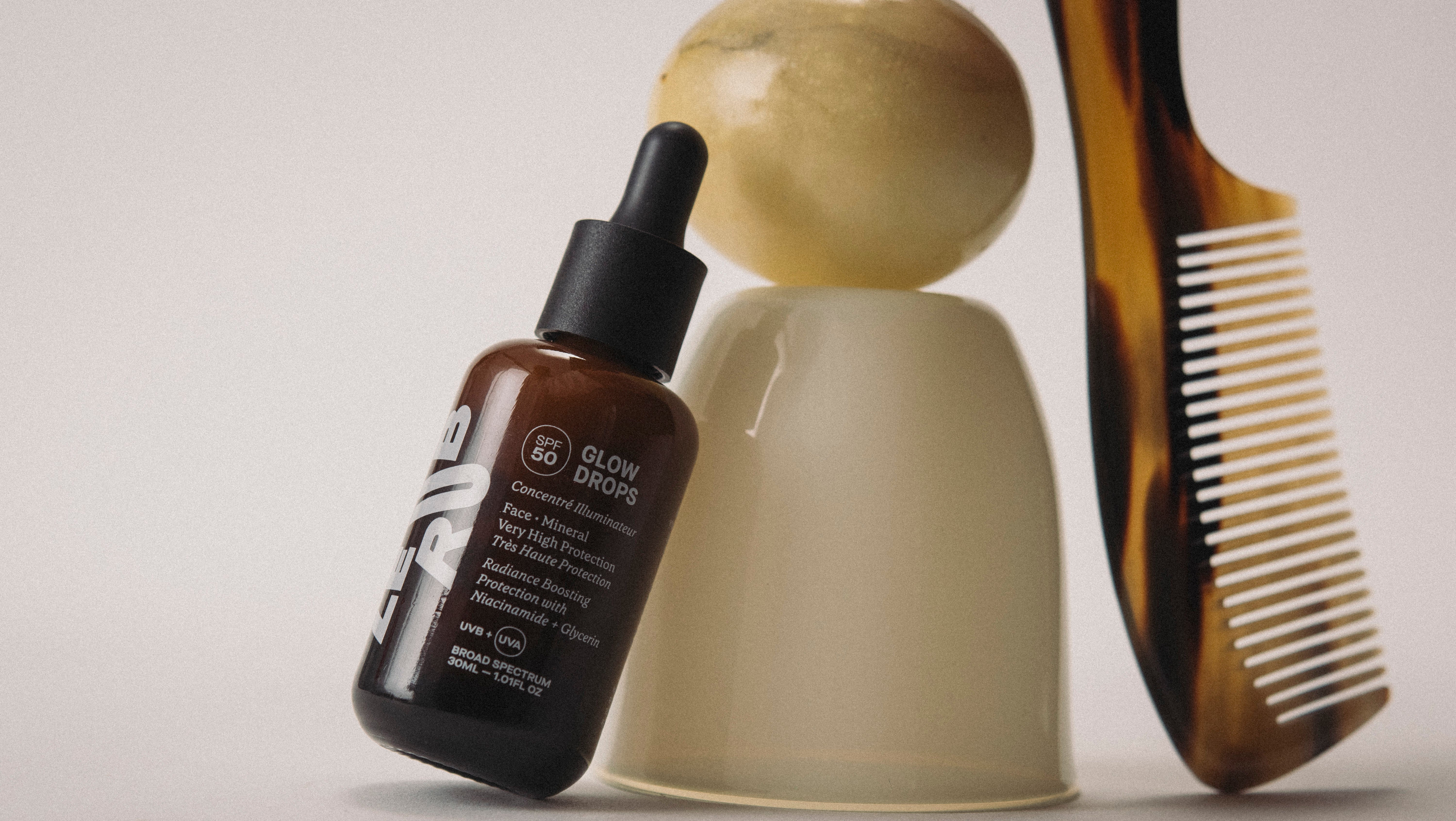 Le Rub Glow Drops SPF50 with comb and decorative objects on an off white studio background