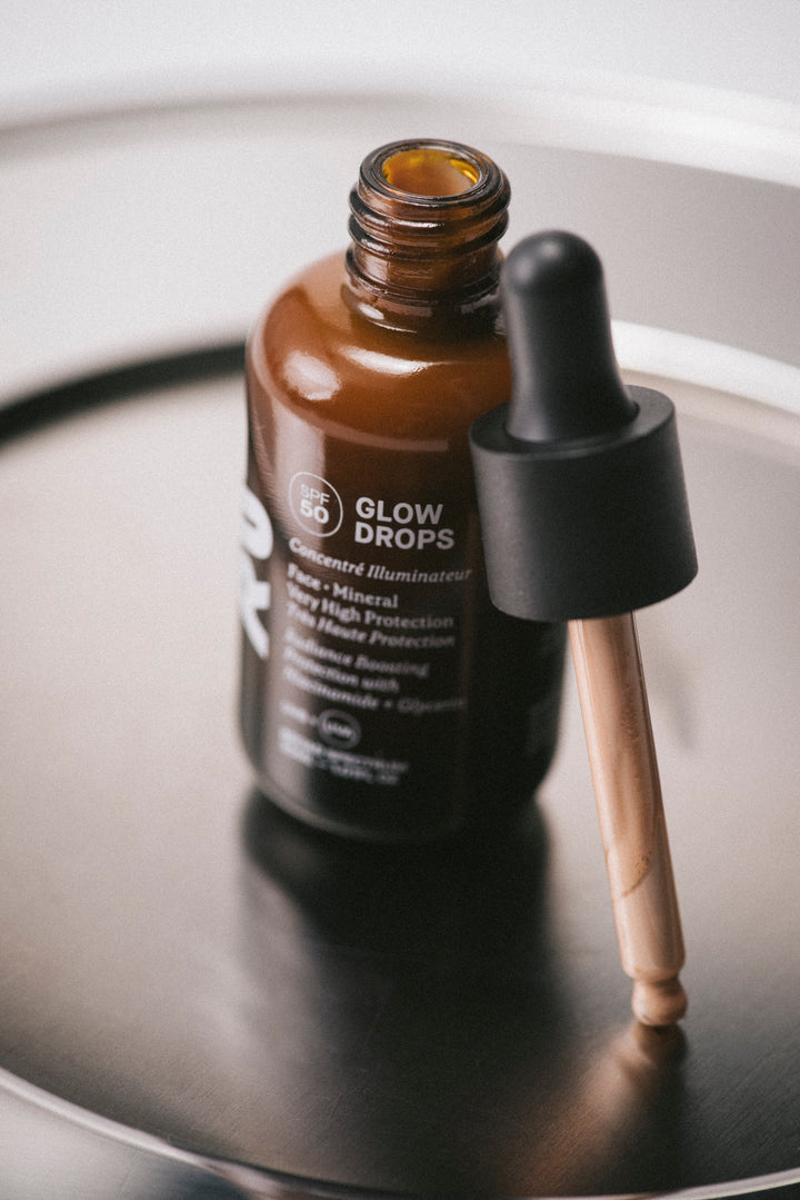 Brown bottle with dropper labeled 'Glow Drops' on a reflective surface. Pipette on the side.